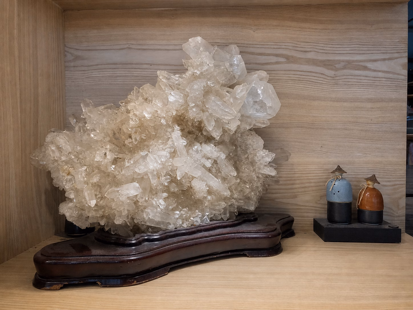 木櫃中的大型透明至乳白色晶簇，置於深色木座旁，右側有兩件小擺飾
